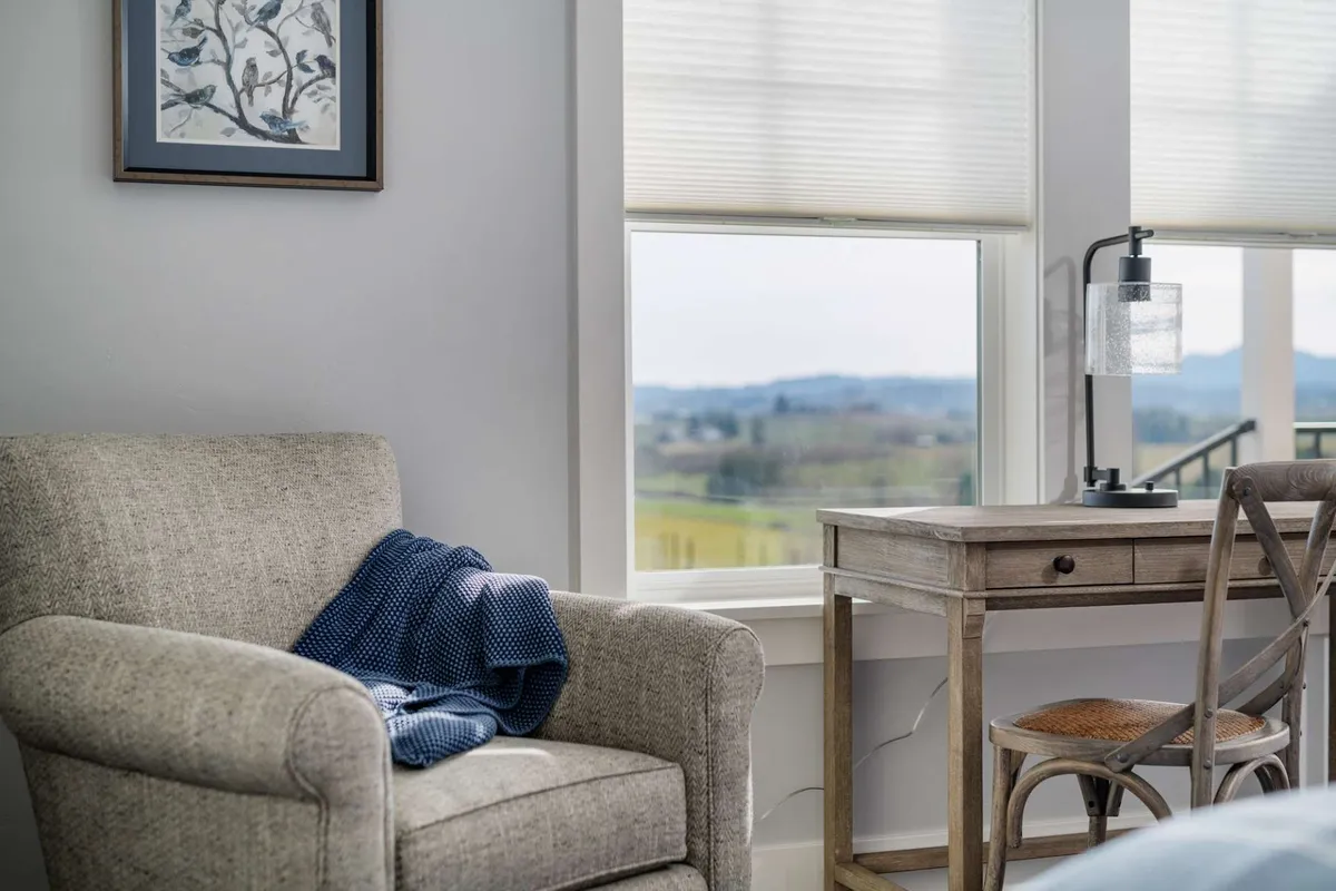 A cozy armchair is positioned beside a small desk and lamp, with a window offering a clear view of the outdoors. The natural light streaming in invites thoughts of the landscape just beyond the glass, hinting at the surrounding countryside and the potential for adventure beyond the home.