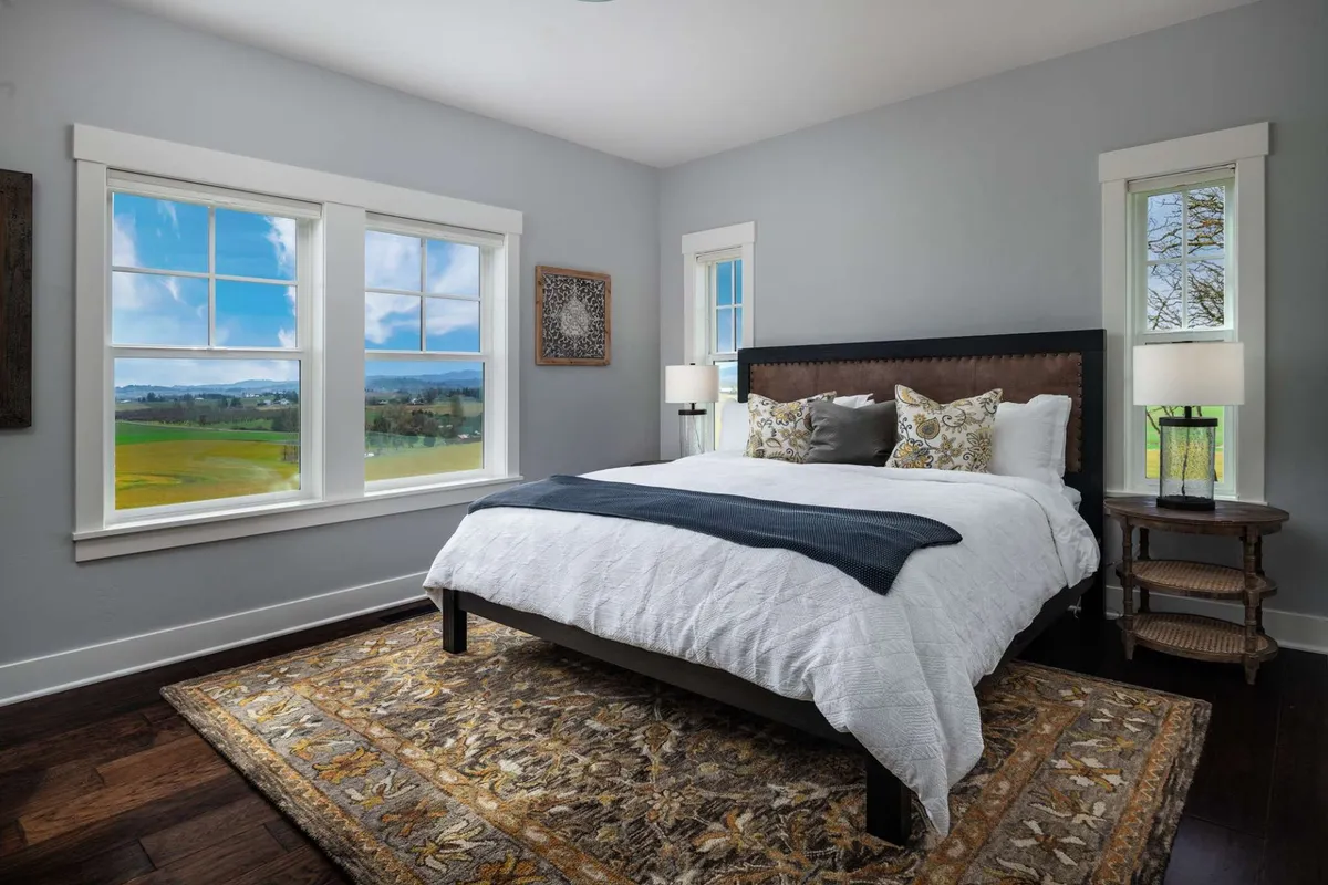 A serene bedroom scene featuring a comfortable bed, window views that let in abundant light, decorative artwork, and a patterned area rug. The overall vibe is tranquil, inviting guests to unwind and rejuvenate.