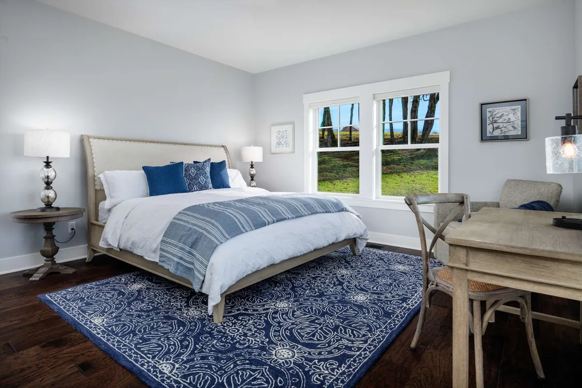 This image highlights a spacious king bedroom featuring a large bed adorned with decorative pillows, stylish bedside lamps, a cozy rug, and a desk and chair positioned by the window. Natural light fills the room, enhancing a serene and restful ambiance.