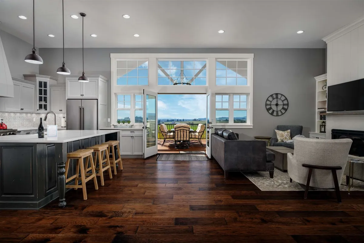 The image showcases a spacious, sunlit kitchen with a large island perfect for gathering, open doors leading to a patio, and sweeping views of the lush Willamette Valley. The inviting interior flows seamlessly to the outdoors, setting the stage for both cozy indoor moments and lively al fresco entertaining.