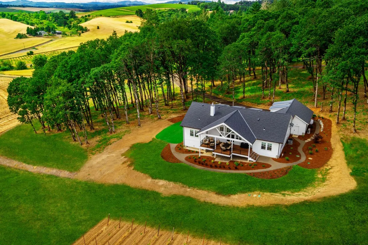 The image showcases the farmhouse nestled among lush trees and open fields, with landscaped gardens in the foreground. The broad view emphasizes the serenity and natural beauty typical of Willamette Valley, while also suggesting outdoor activities and gatherings.