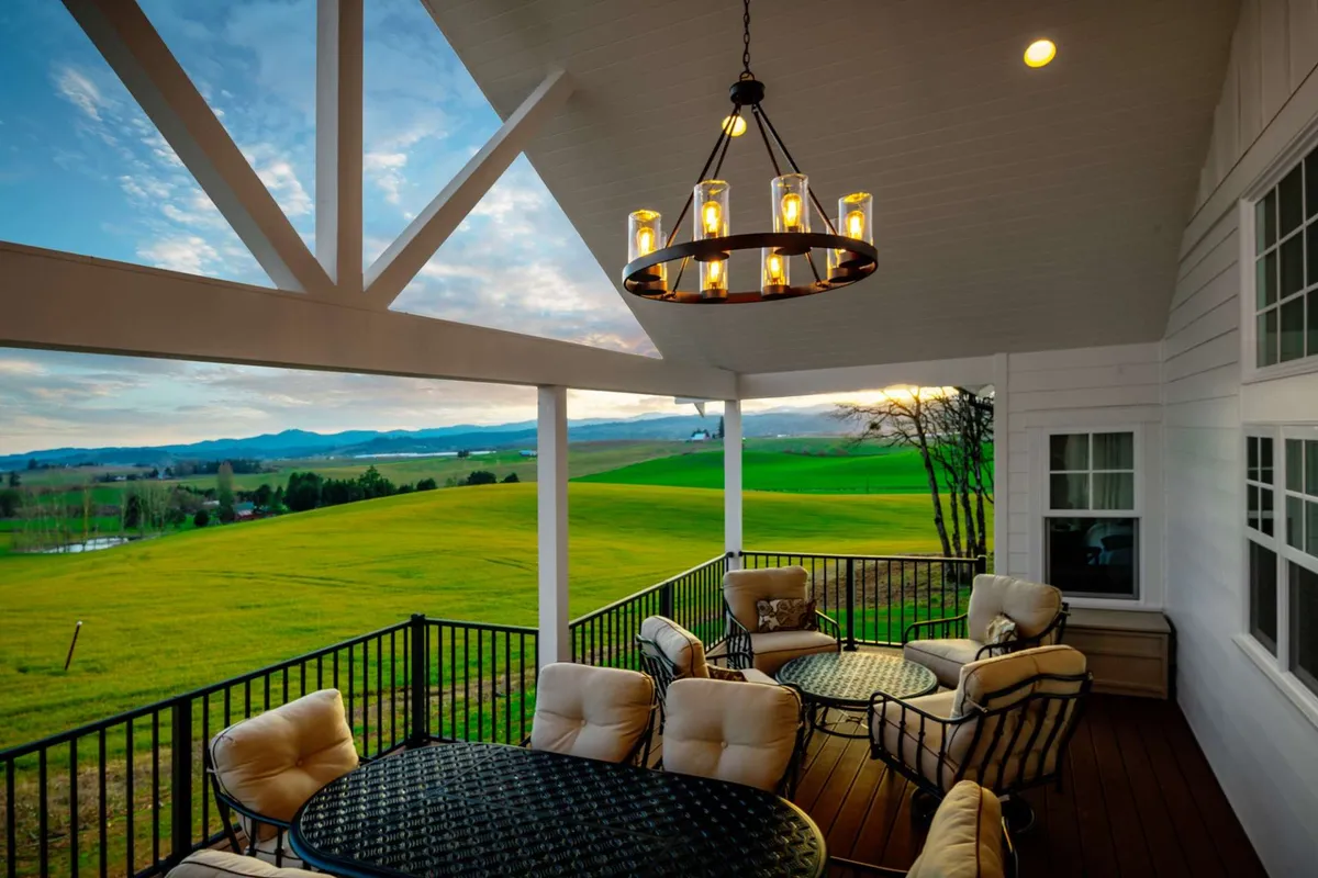 An expansive outdoor deck is styled with elegant outdoor furniture beneath a chandelier, overlooking lush green fields and distant mountains. The seating area is inviting and arranged for conversation or relaxation as the sun sets beyond the horizon.